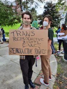 image of a person at a rally holding a cardboard sign that reads "trans rights are reproductive rights"